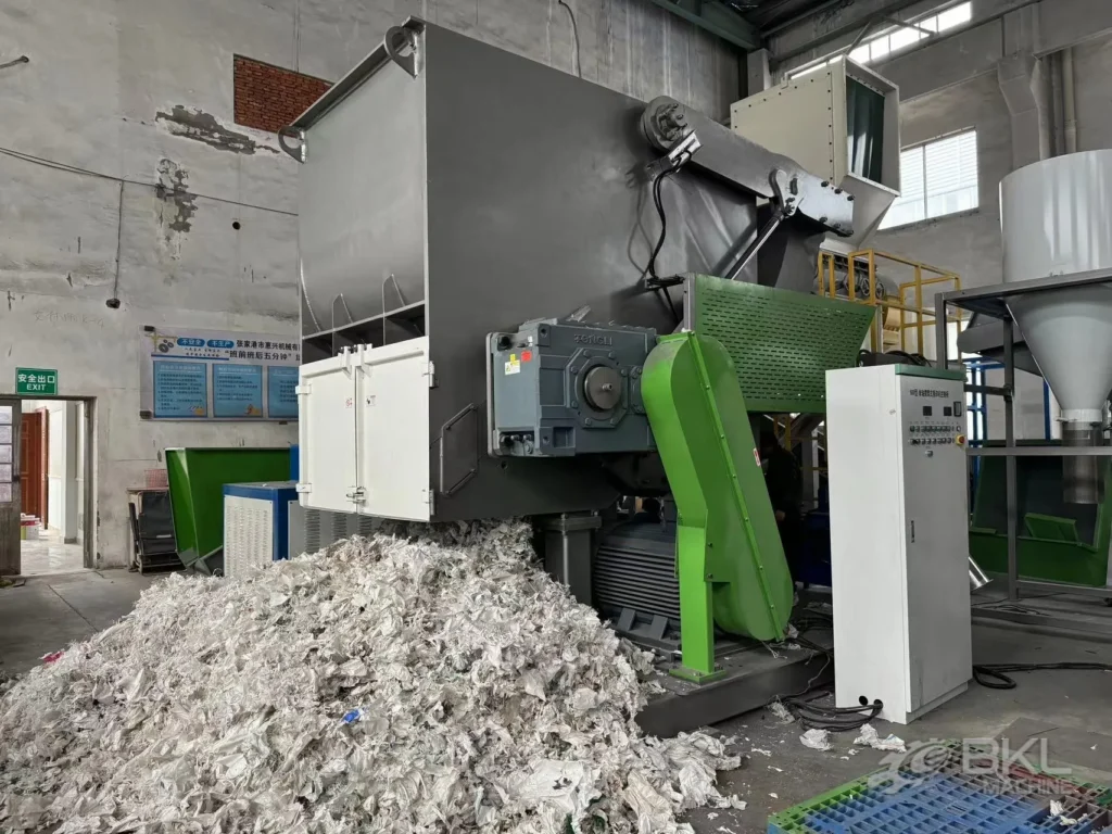 Single-shaft shredder in operation with a pile of shredded white film flakes and a control cabinet