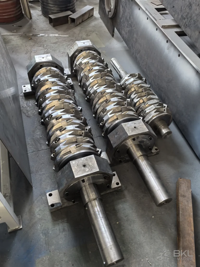 Two single-shaft shredder rotors on the workshop floor with helically bolted blades and machined journals