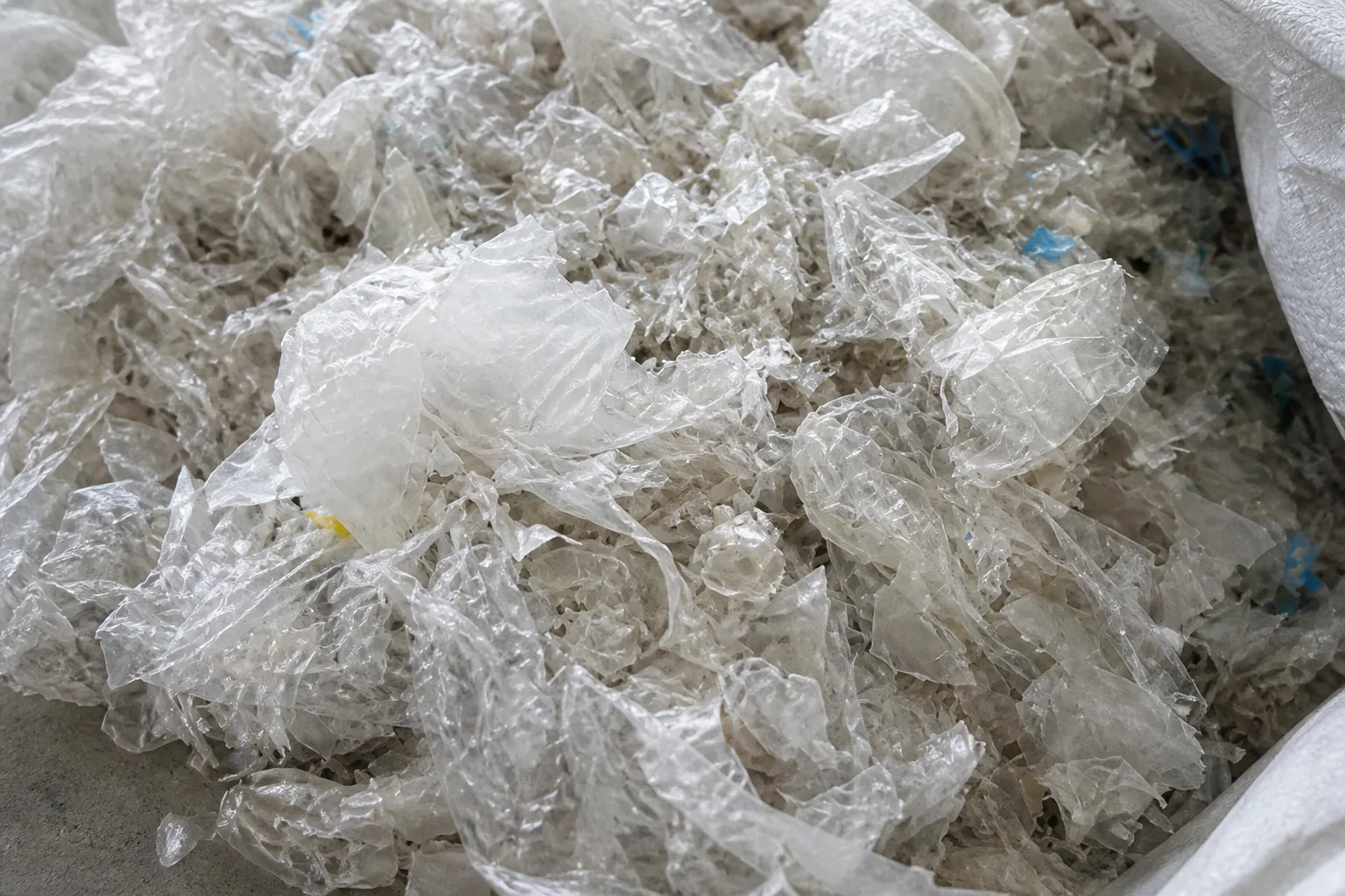 Heap of washed translucent white film flakes, curled and crumpled, piled inside a woven bulk bag beside a strip of grey concrete floor.
