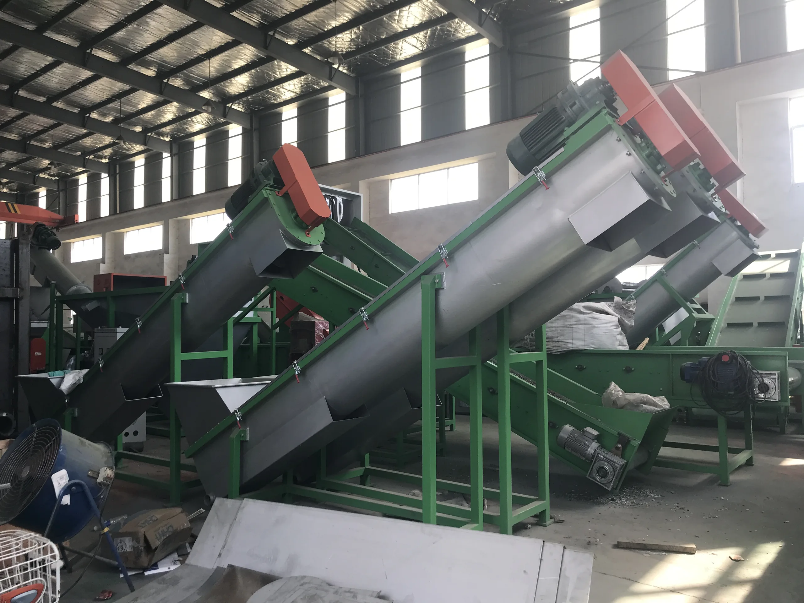 Inclined screw loaders feeding green washing machines inside a plant, with loose material lying on the floor between them.