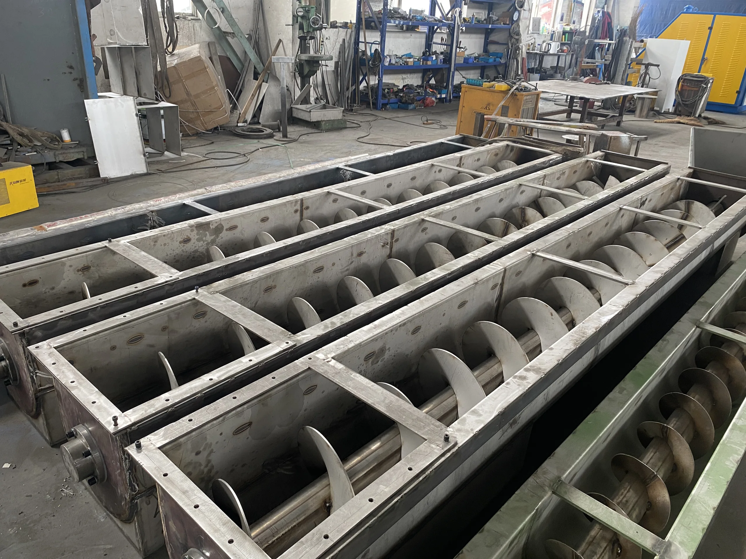 Two long open stainless steel screw conveyor troughs with helical augers inside, lined up outdoors before installation.