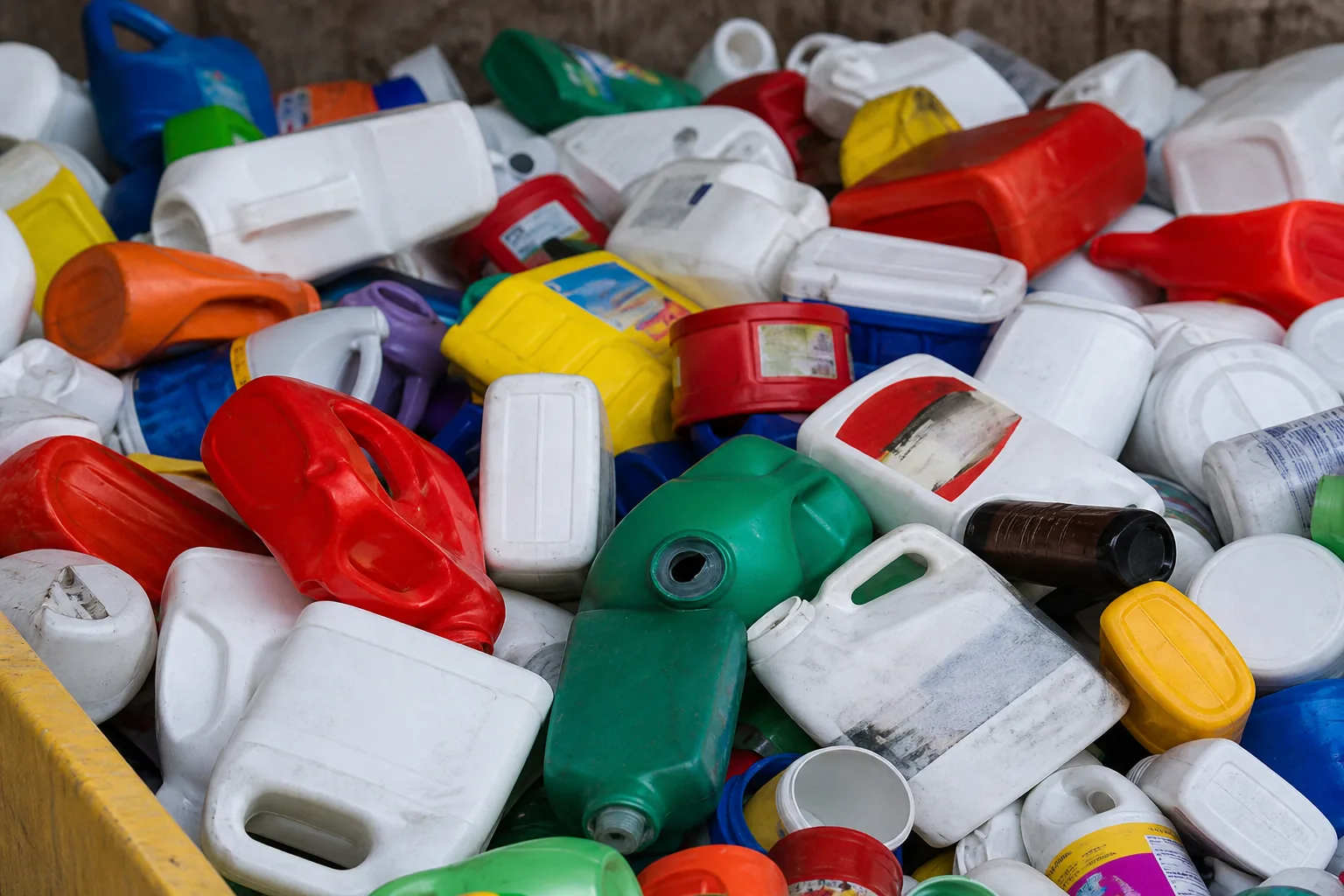 Heap of mixed post-consumer rigid plastic bottles, jugs and canisters in many colours, dented from handling and still carrying labels.
