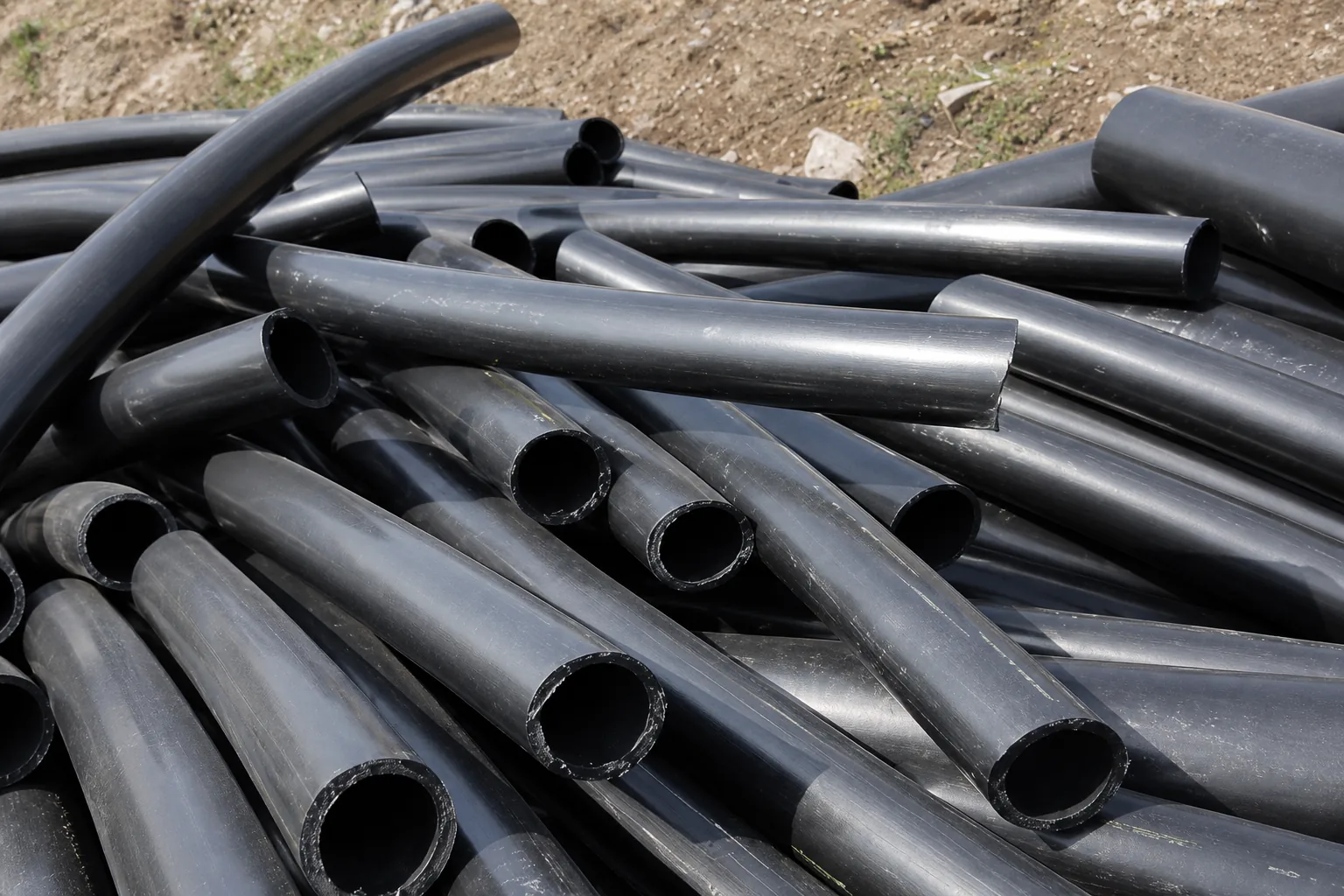 Loose heap of thick-walled black plastic pipe offcuts lying on bare ground, their cut circular ends facing the camera.