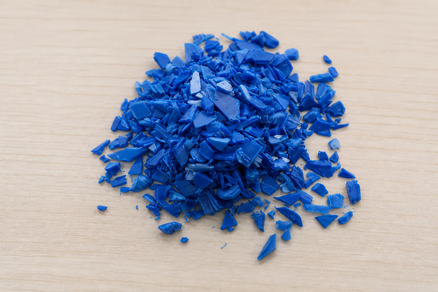 Small mound of washed blue rigid plastic regrind, hard angular chips with sharp fractured edges, on a pale wooden bench.