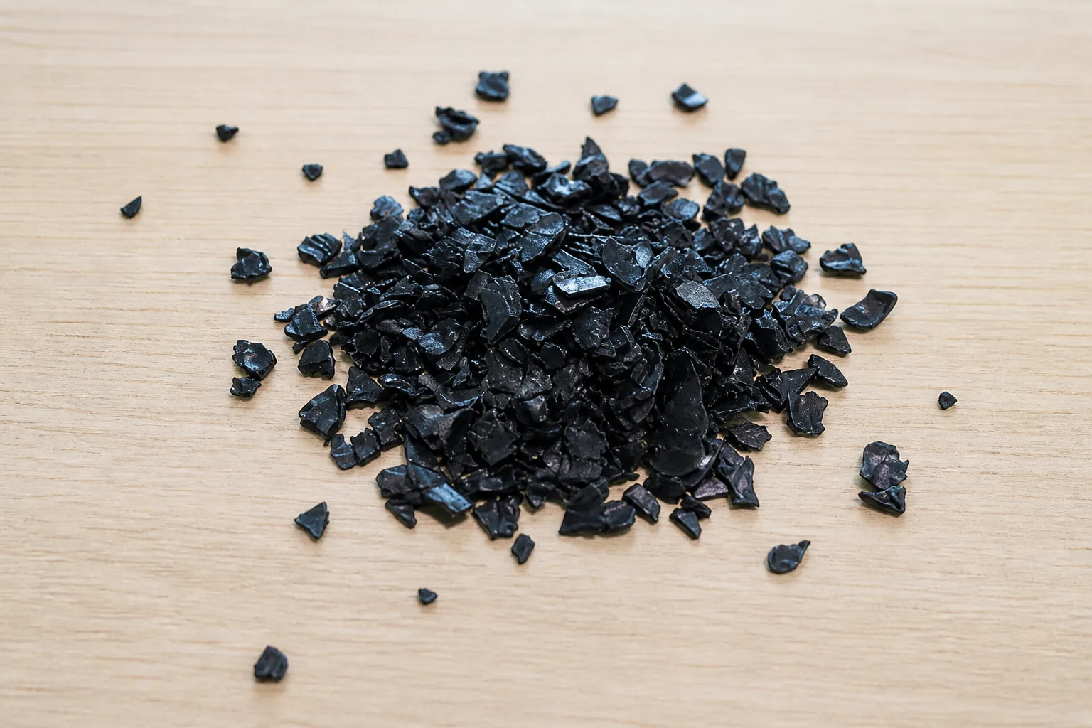 Loose mound of washed black rigid plastic regrind with the broken edges catching the light against a pale wooden bench.